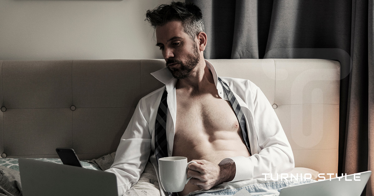 Handsome man with his shirt open working on his computer and holding a mug loose tie around his neck