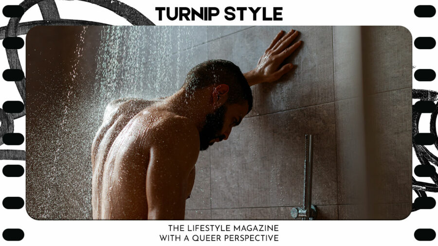 A man standing in the shower, water falling on to his back and head, his left arm is up on the wall as he looks down to the floor